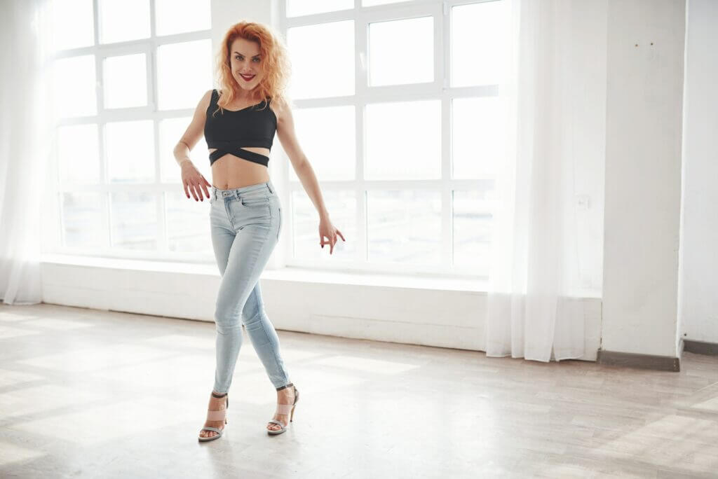 In high heels. Attractive redhead woman posing in the spacey room near the window
